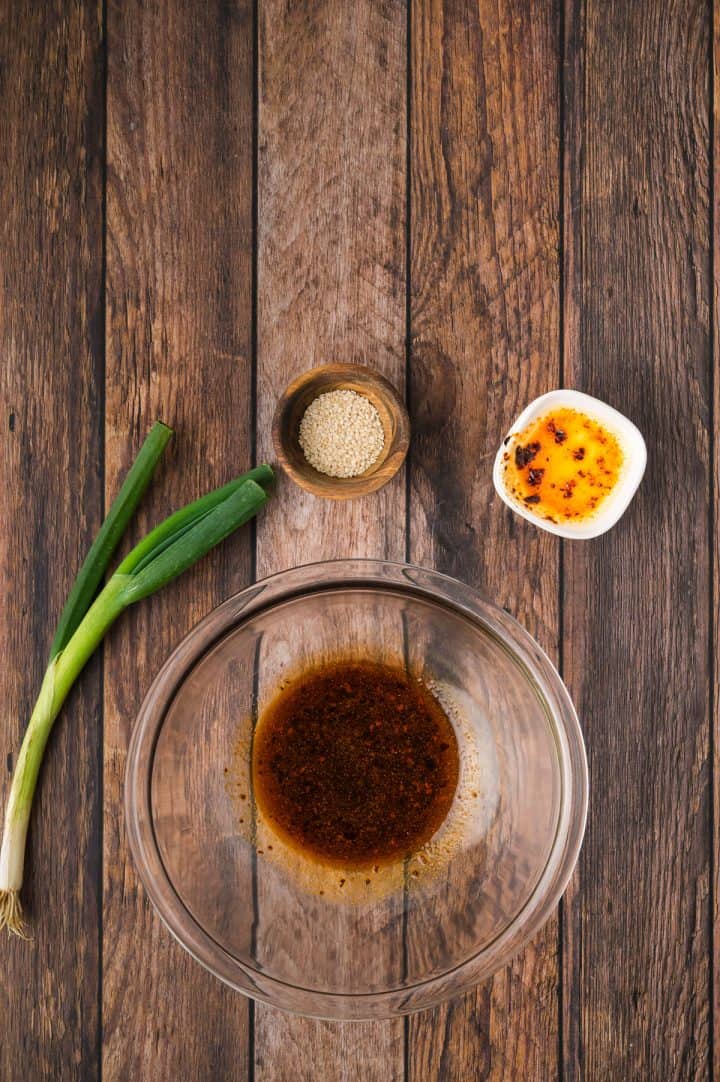 Glass bowl with sauce, green onions, sesame seeds, and chili oil on a wooden table.
