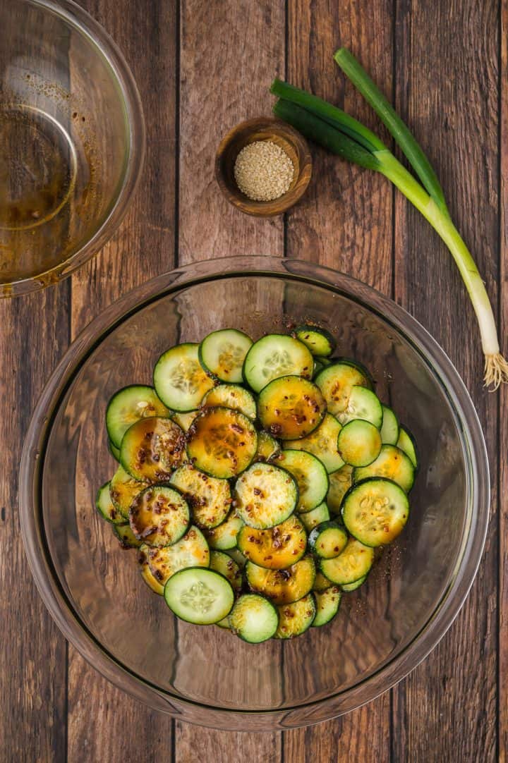 Sliced cucumbers in a glass bowl with sauce, next to sesame seeds and a green onion on a wooden table.
