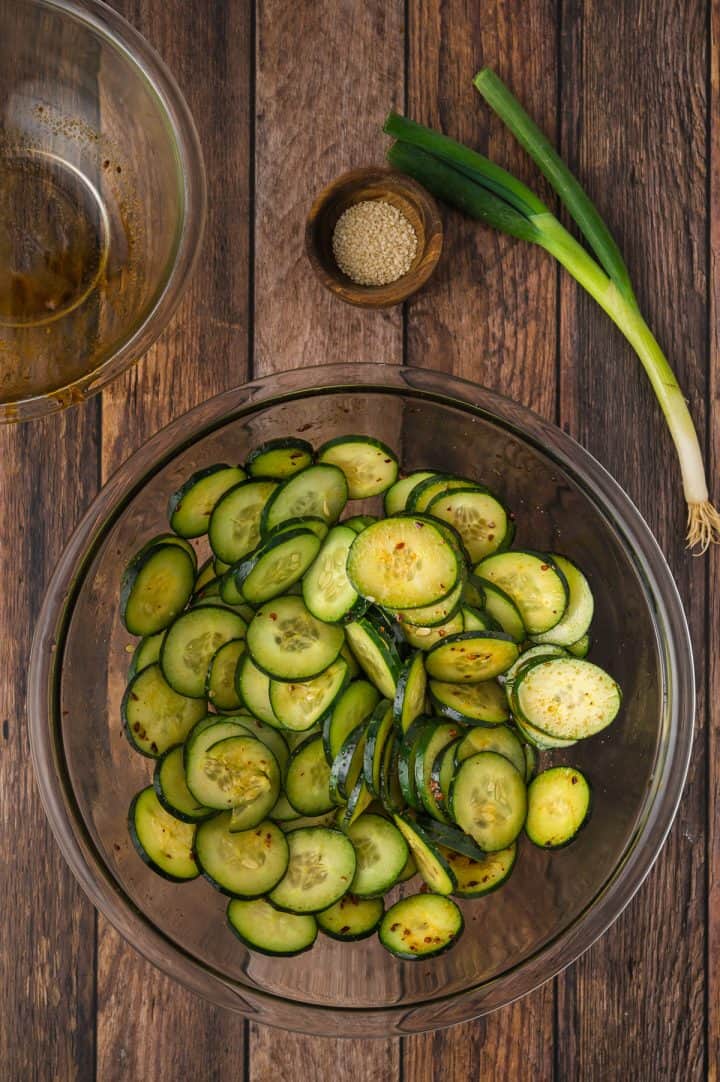 Sliced cucumber in a glass bowl with green onions, sesame seeds, and an empty glass bowl on a wooden table.