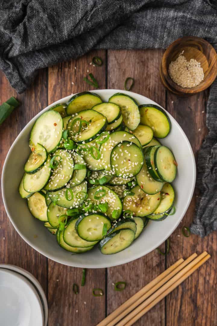 Sliced cucumber salad with sesame seeds in a white bowl, on a wooden table with chopsticks and a gray napkin.