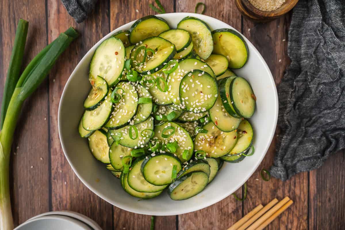 Bowl of sliced cucumber salad with sesame seeds on a wooden table, next to scallions and chopsticks.