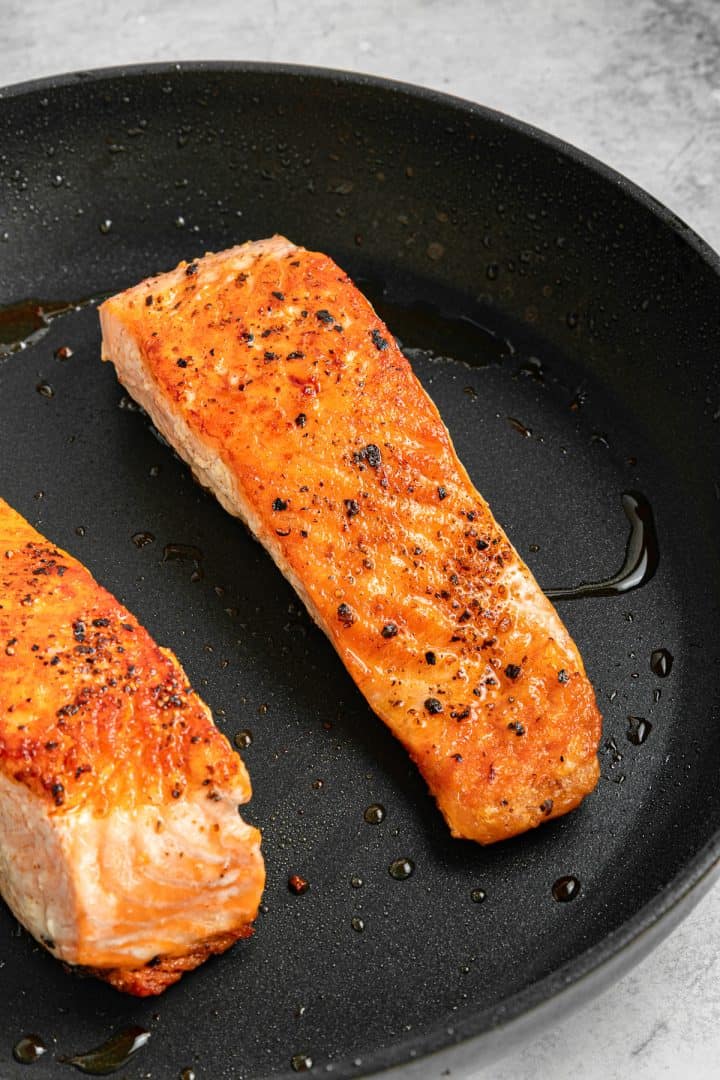 Two seasoned salmon fillets cooking in a black nonstick pan on a gray surface.