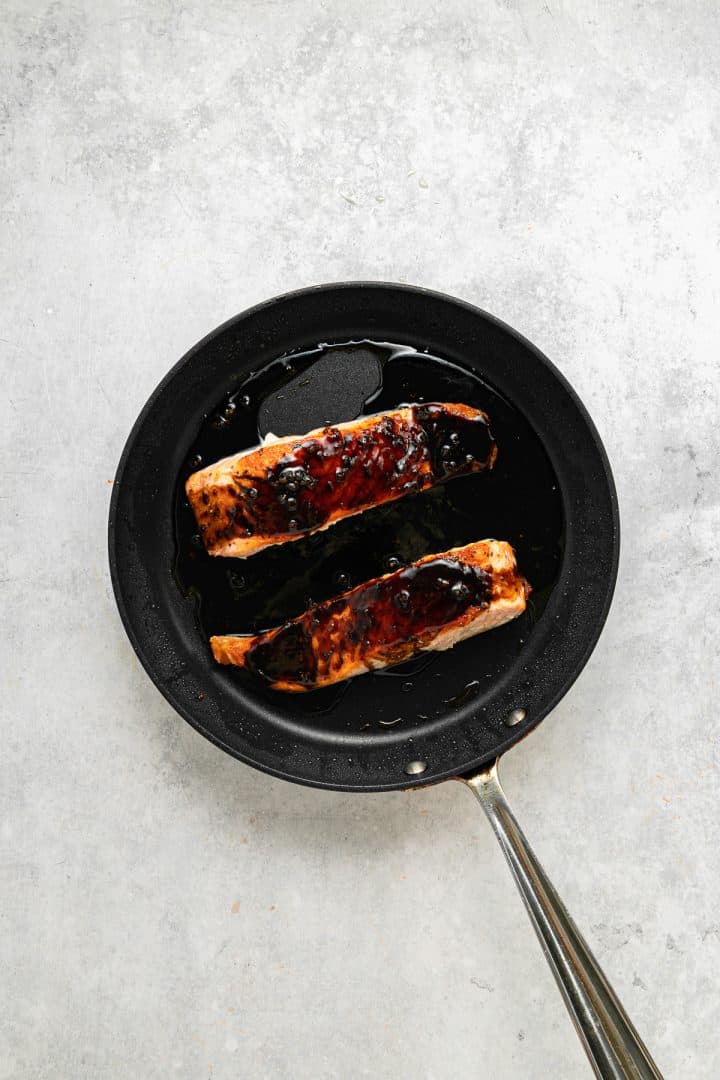 Two pieces of glazed salmon cooking in a black frying pan on a light gray surface.