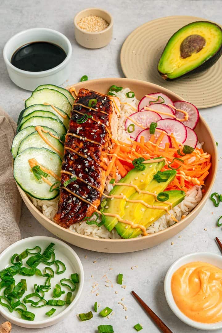 Salmon rice bowl with sliced cucumber, radish, avocado, shredded carrots, and green onions, drizzled with sauce.