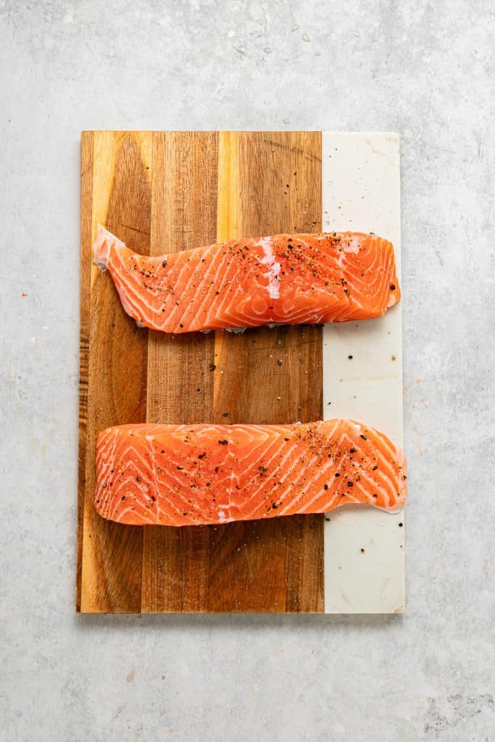 Two raw salmon fillets with pepper on a wooden and marble cutting board, seen from above.