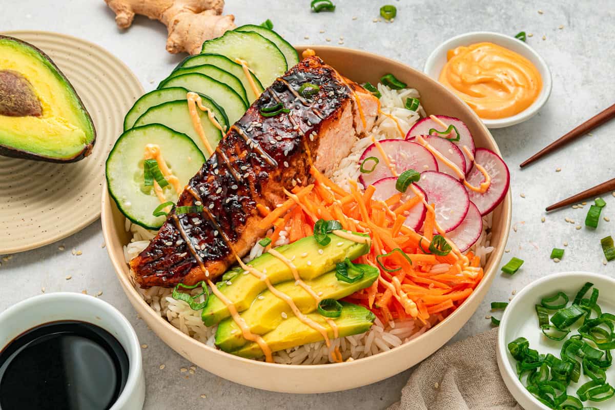 A salmon rice bowl with avocado, cucumber, radish, carrot, sauce, and green onions, served with chopsticks.