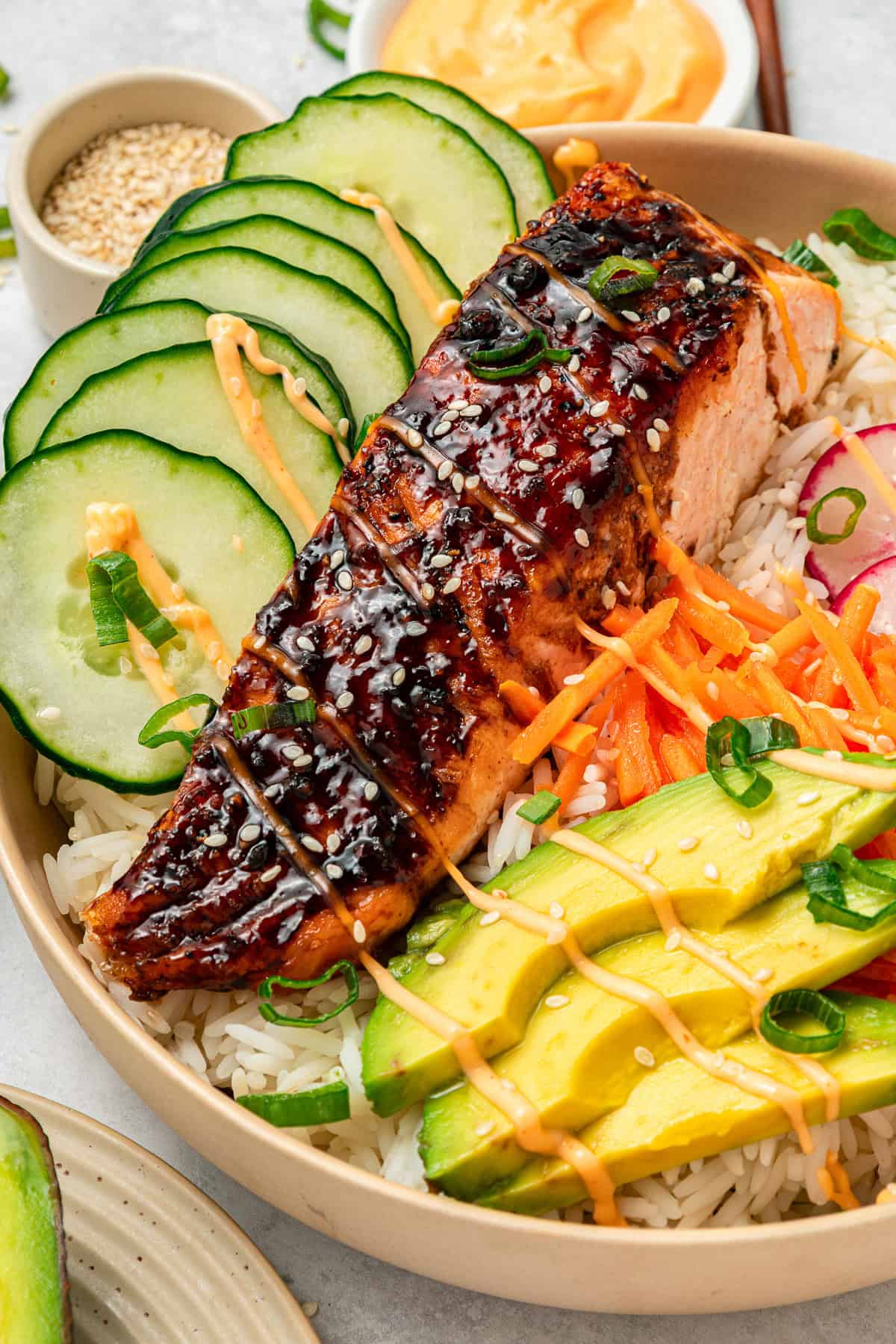 Grilled salmon on rice with sliced cucumber, avocado, shredded carrots, and a drizzle of creamy sauce.