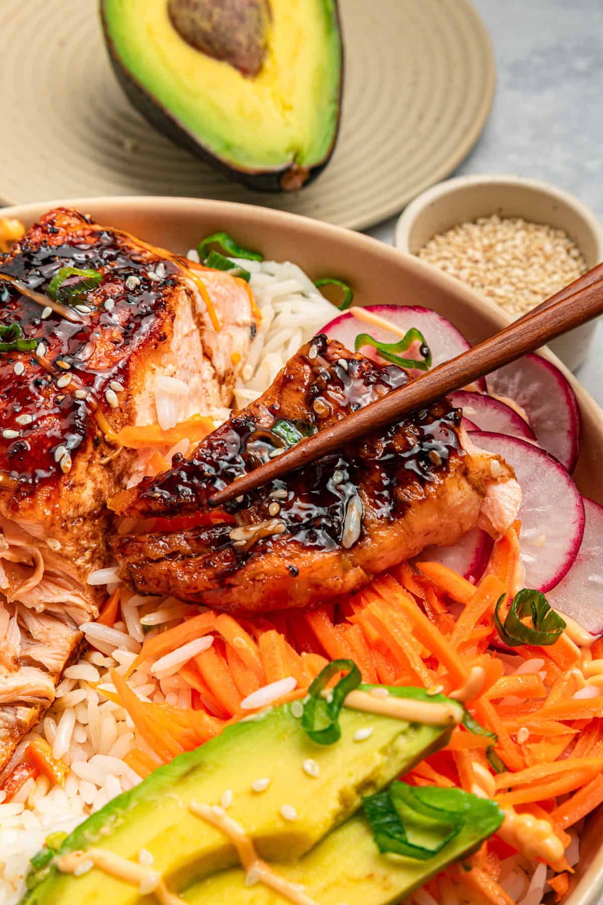 Chopsticks holding glazed salmon over rice, carrots, radish, and avocado slices, topped with sesame seeds.