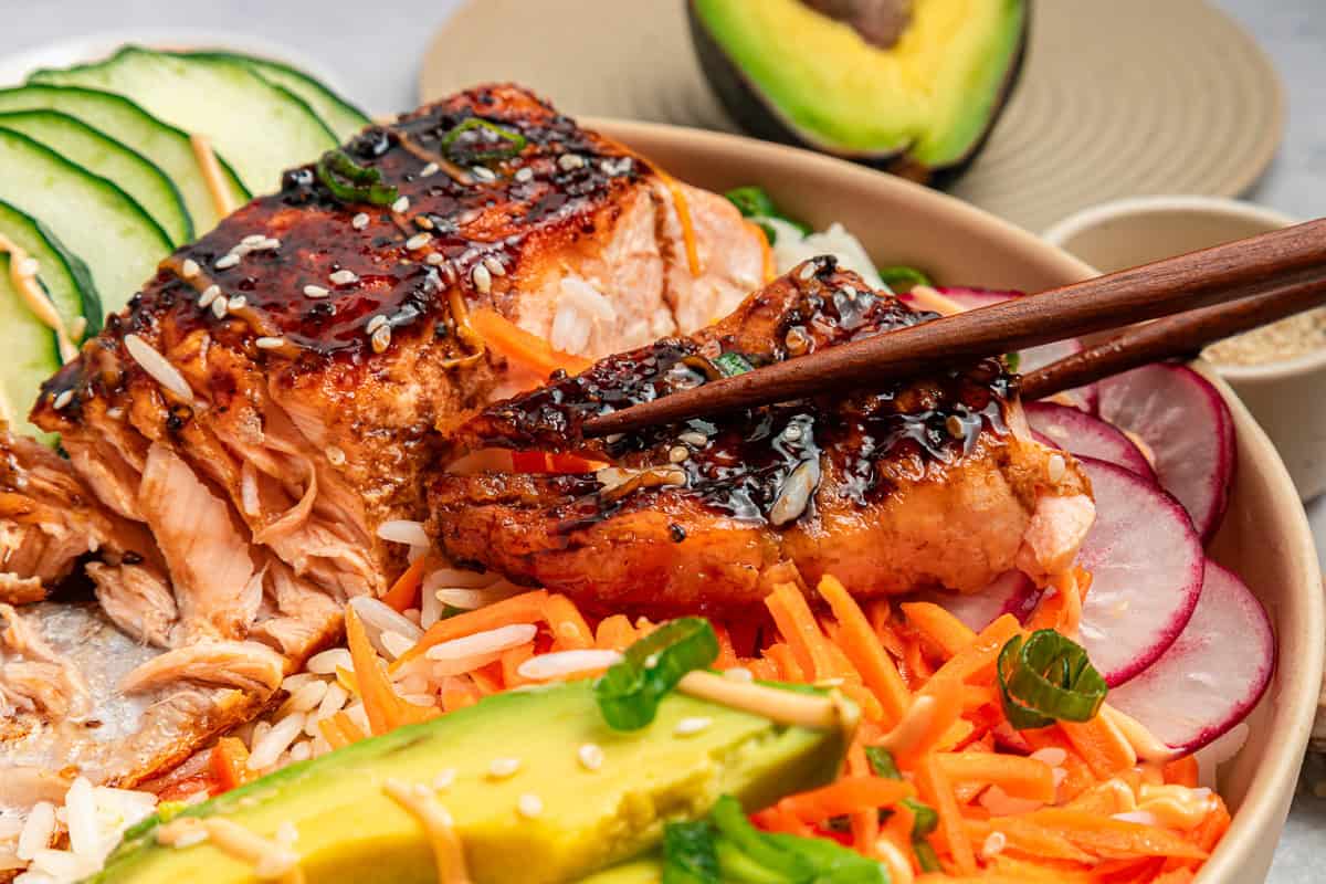 Chopsticks hold glazed salmon over rice, veggies, cucumber, radish, carrot, and avocado slices.