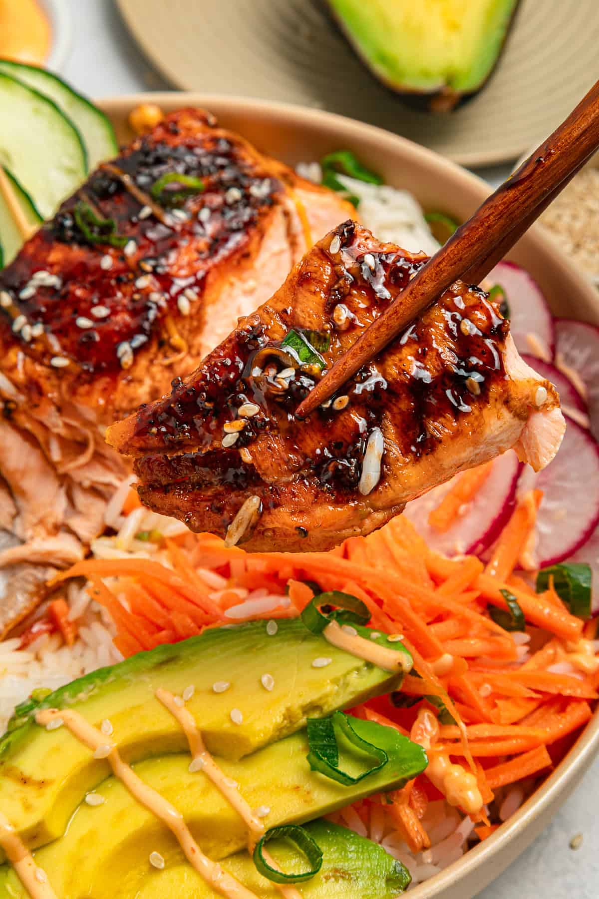 Chopsticks hold glazed salmon over a bowl with rice, avocado, carrots, radish, and sesame seeds.