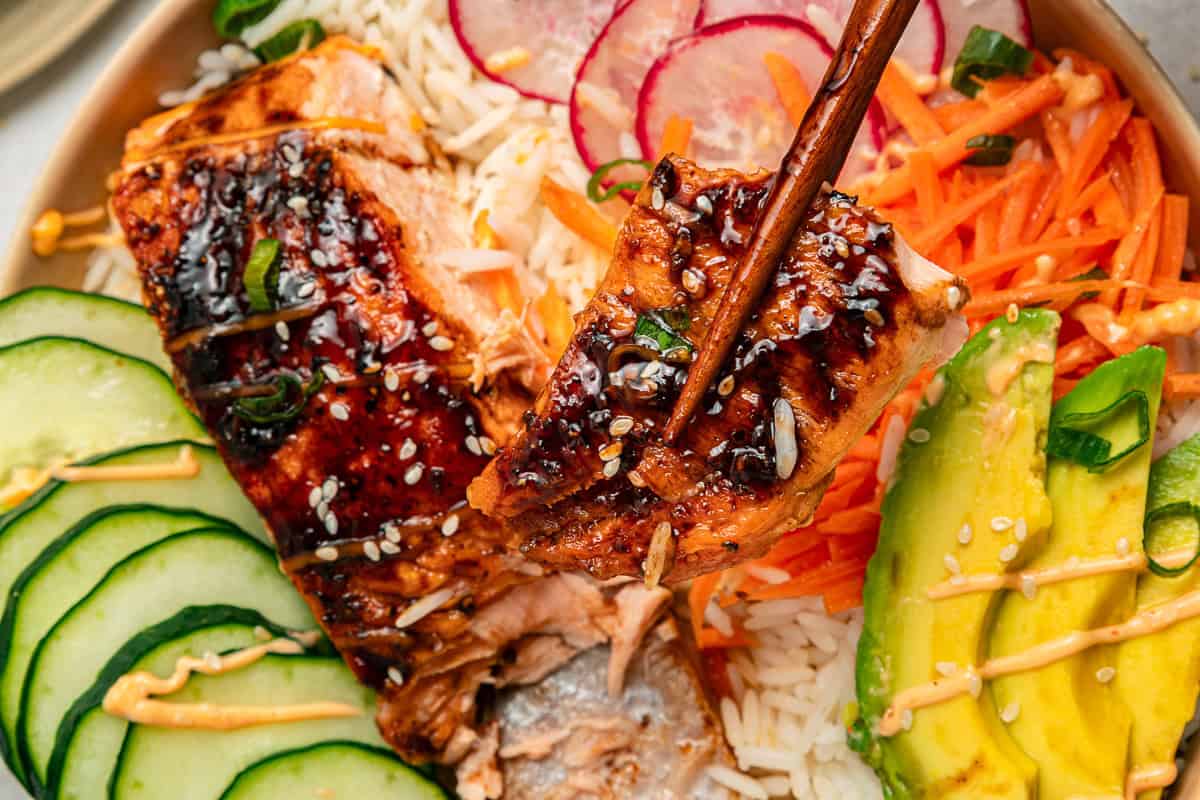 Grilled salmon with rice, sliced cucumber, avocado, carrots, and radishes, topped with sauce and sesame seeds.