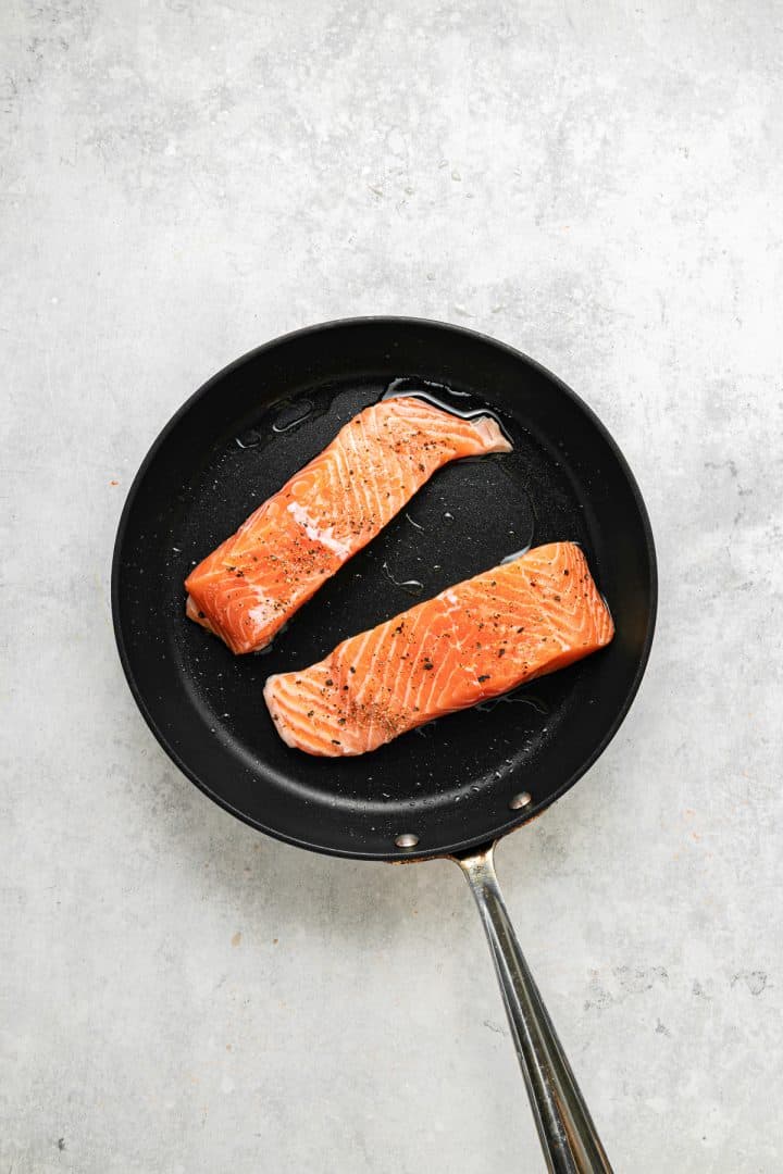 Two raw salmon fillets in a black frying pan on a light gray textured surface, viewed from above.