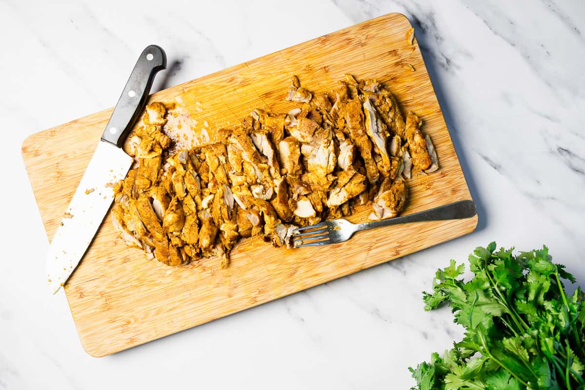 Sliced cooked chicken with a knife and fork on a cutting board, cilantro nearby on a marble surface.