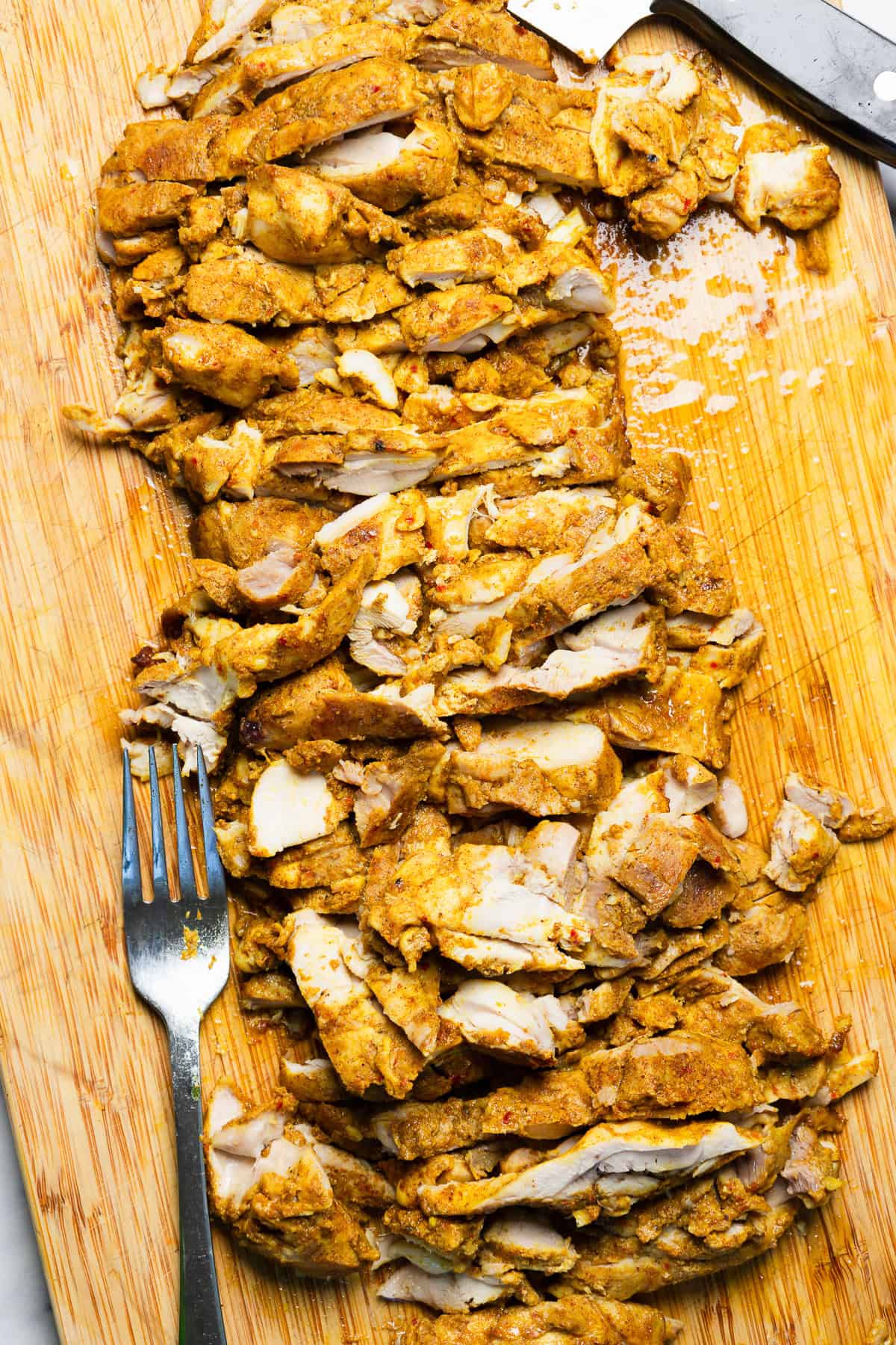 Sliced spiced chicken on a wooden cutting board with a fork and knife nearby.