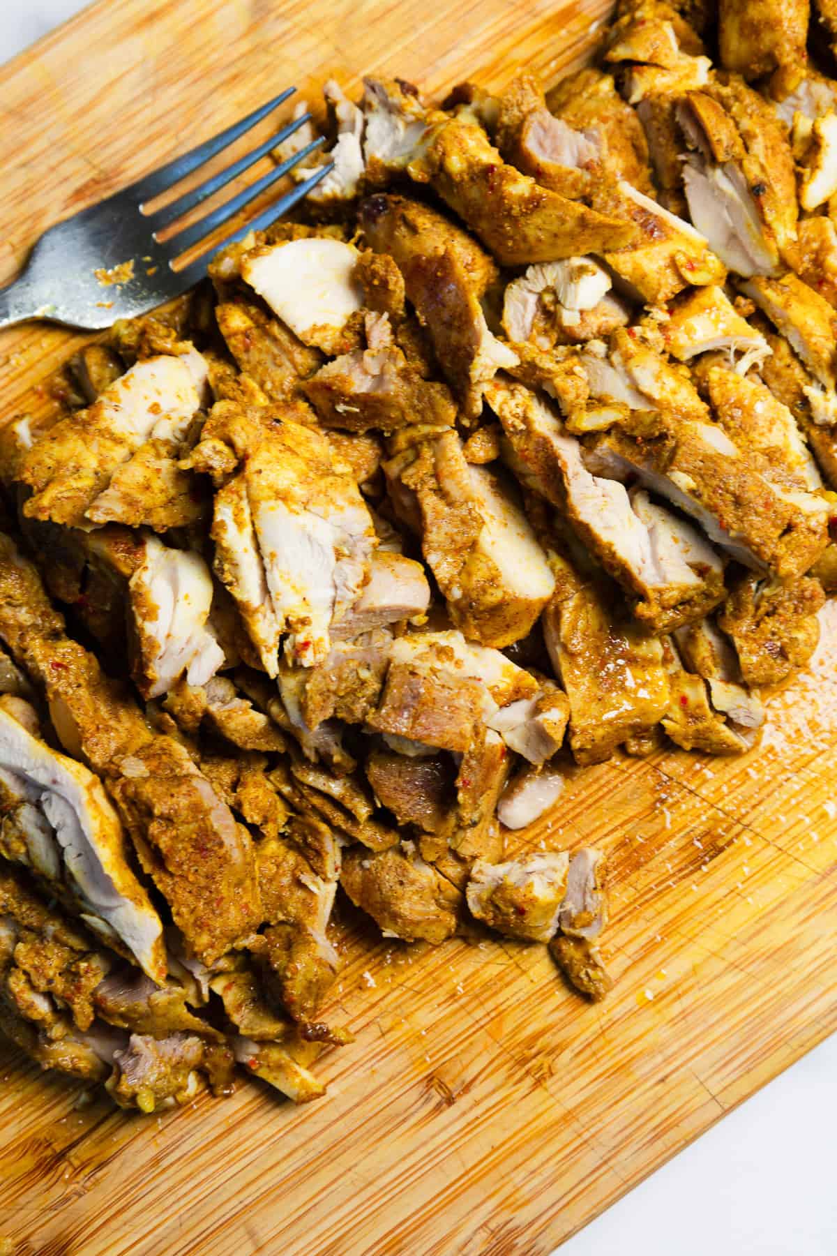 Sliced cooked chicken seasoned with spices on a wooden cutting board with a metal fork nearby.