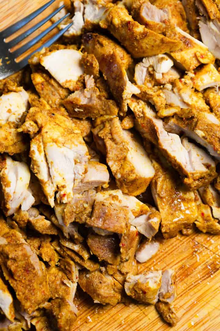 Sliced seasoned cooked chicken pieces on a wooden cutting board with a metal fork nearby.