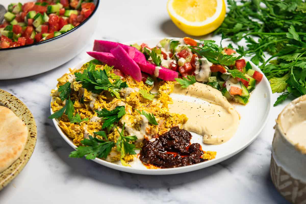 Plate with shredded chicken, hummus, salad, pickled veggies, hot sauce, and fresh parsley on marble table.