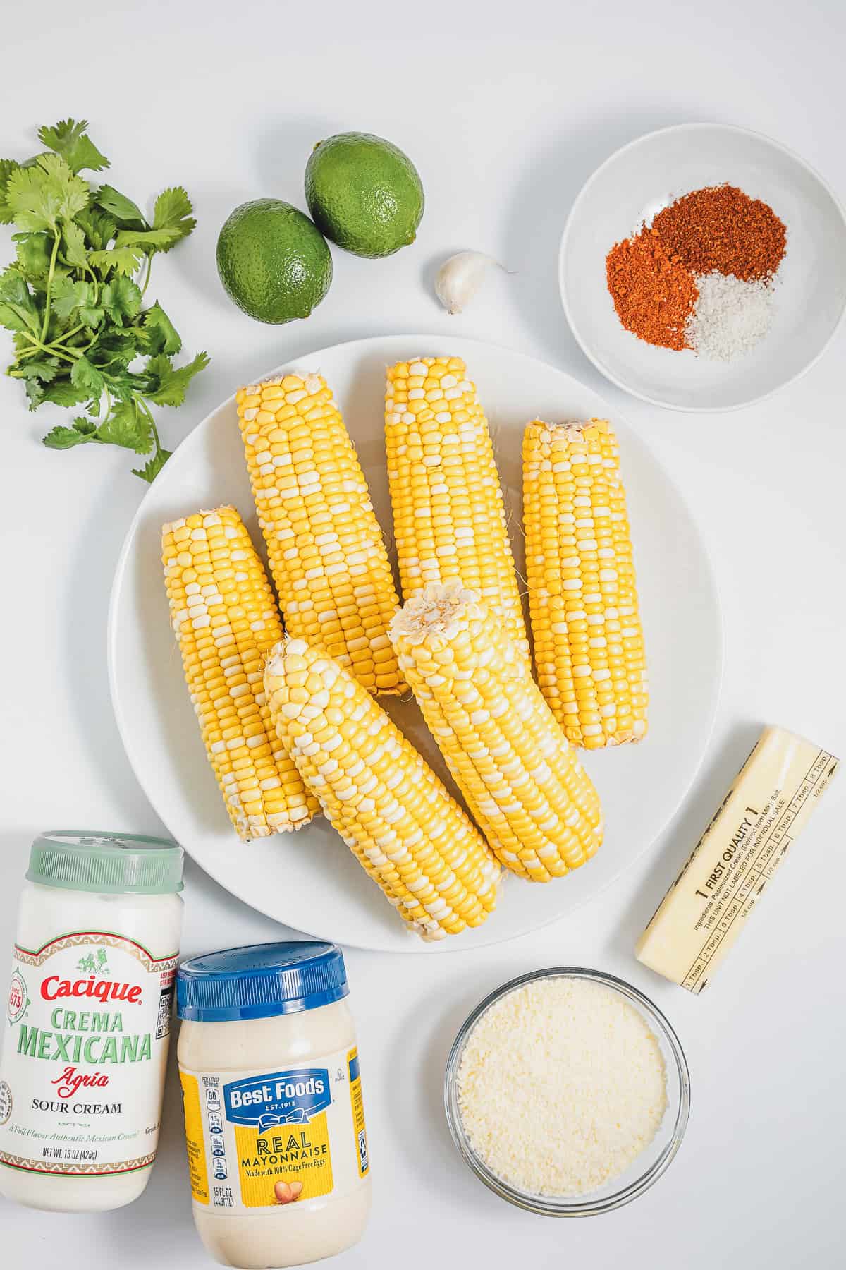 Six corn cobs on a plate with limes, cilantro, garlic, spices, butter, sour cream, mayo, and cheese around—perfect for making authentic Mexican street corn elotes.
