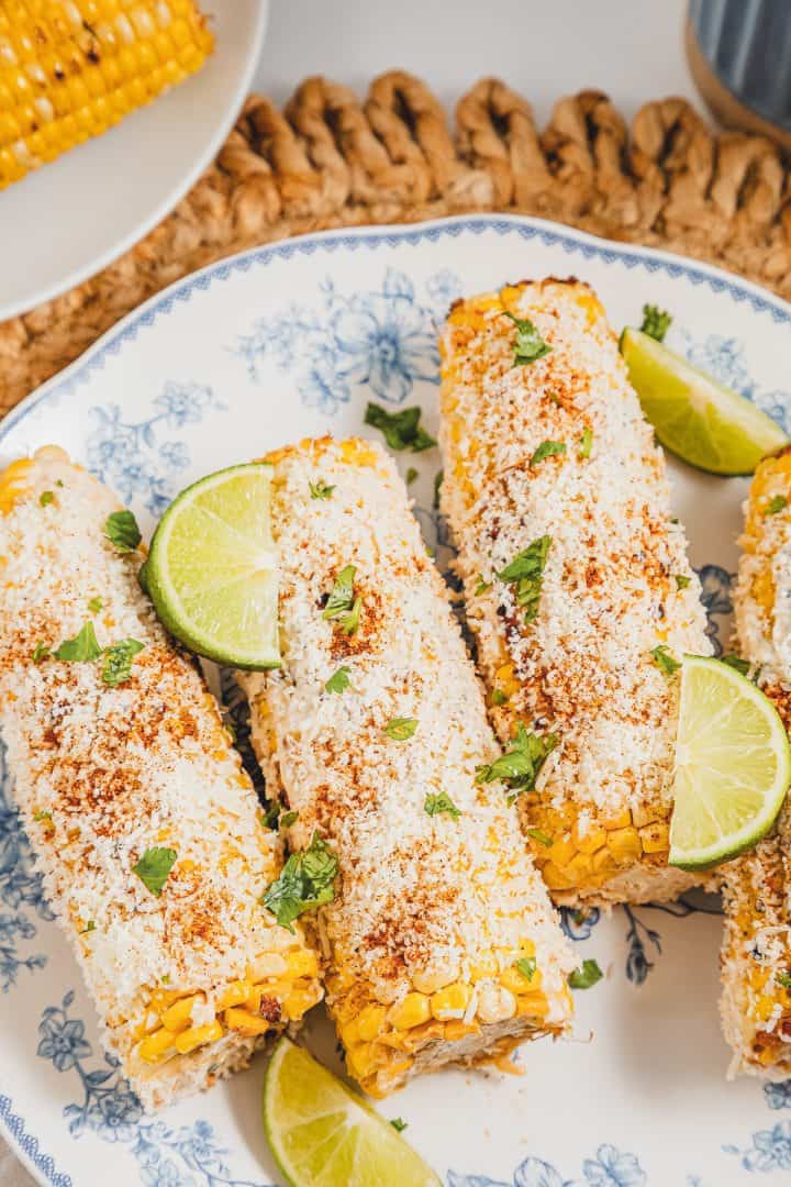 Four Mexican street corn elotes topped with cheese, chili powder, and cilantro, garnished with lime wedges on a plate.