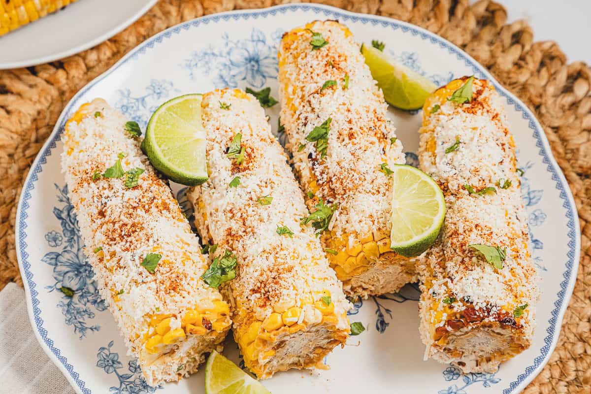 Four delicious mexican street corn elotes on a plate, generously topped with cheese, herbs, and lime wedges.
