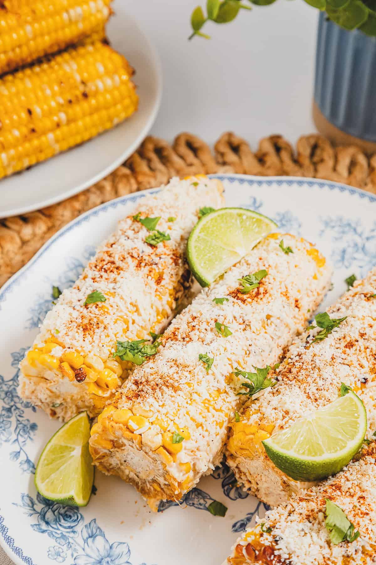 Three Mexican street corns, also known as elotes, topped with cheese, chili powder, cilantro, and lime wedges on a blue-rimmed plate.