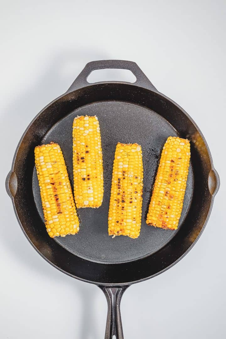 Four grilled corn cobs in a black cast iron skillet on a white background, inspired by Mexican street corn elotes.