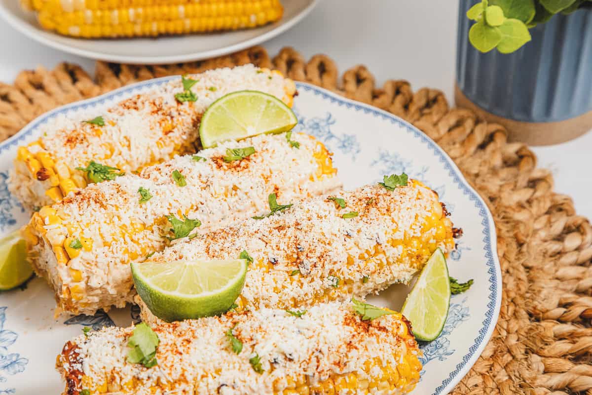 Four mexican street corn elotes sprinkled with cheese, chili powder, and lime wedges on a decorative plate, with a plant nearby.