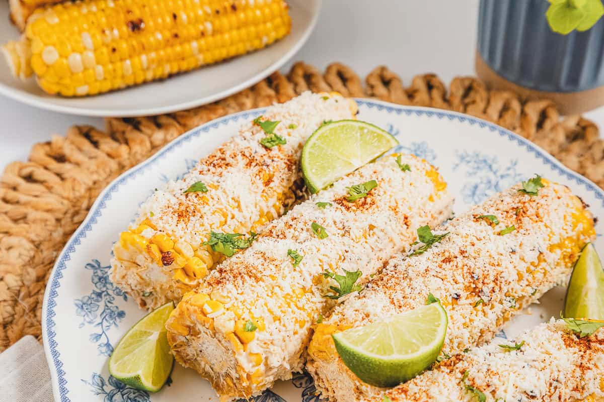 Three ears of Mexican street corn elotes topped with cheese, chili powder, and lime wedges on a patterned plate.