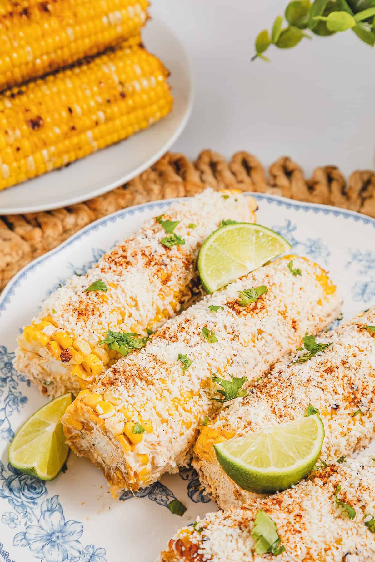 Three ears of Mexican street corn elotes garnished with cheese, chili powder, cilantro, and lime wedges on a plate.