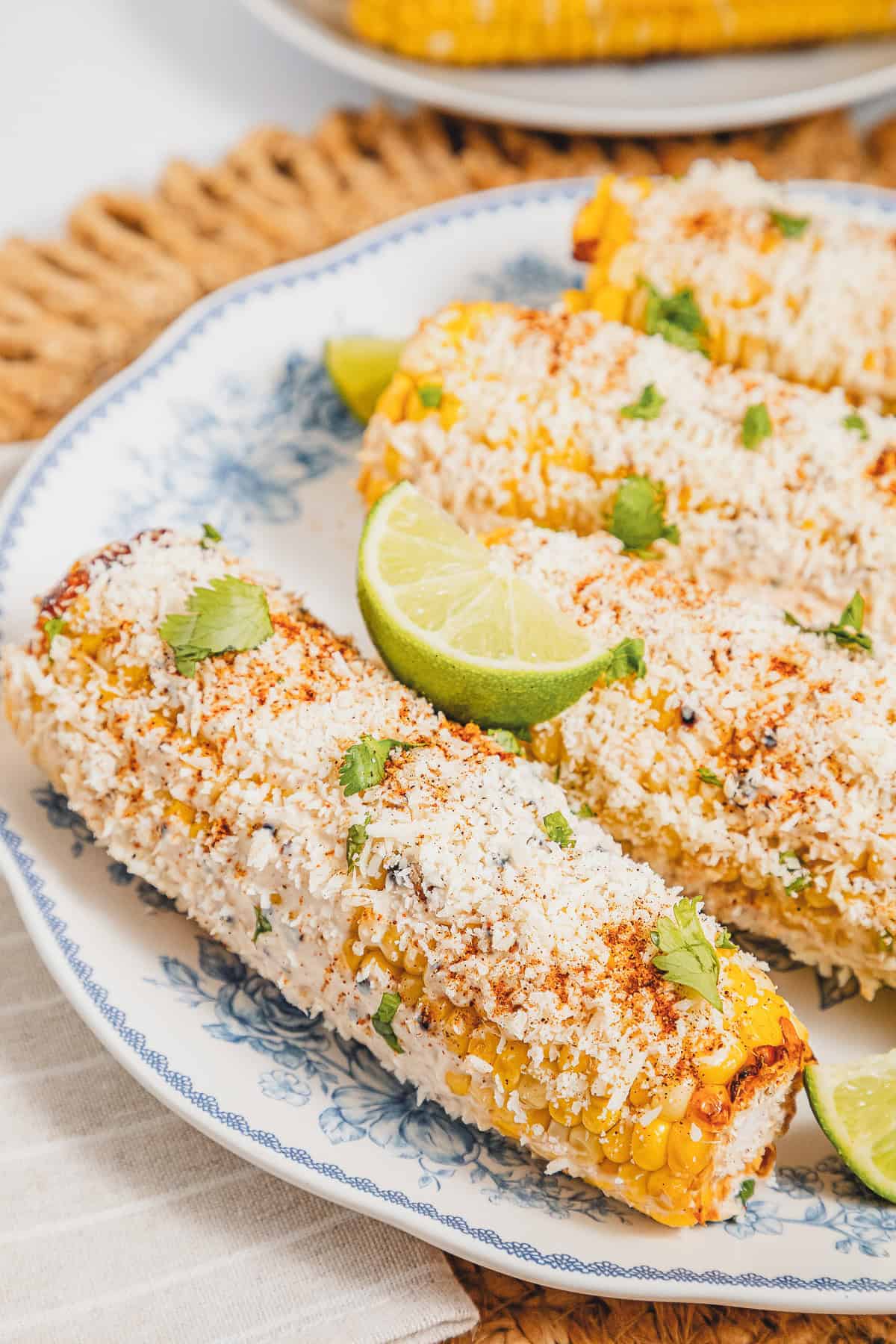Plate of Mexican street corn elotes topped with cheese, spices, cilantro, and lime wedges on a blue floral plate.