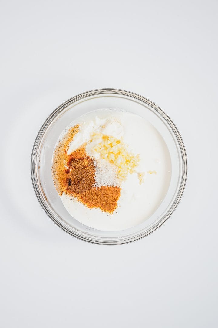 A glass bowl with yogurt, spices, salt, and minced garlic on a white background-perfect for making a creamy sauce inspired by Mexican street corn elotes.