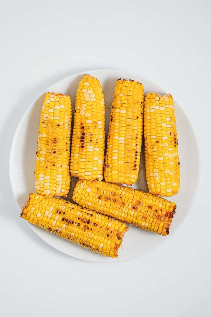 Six grilled corn on the cob pieces, inspired by Mexican street corn elotes, are artfully arranged on a white plate and viewed from above.