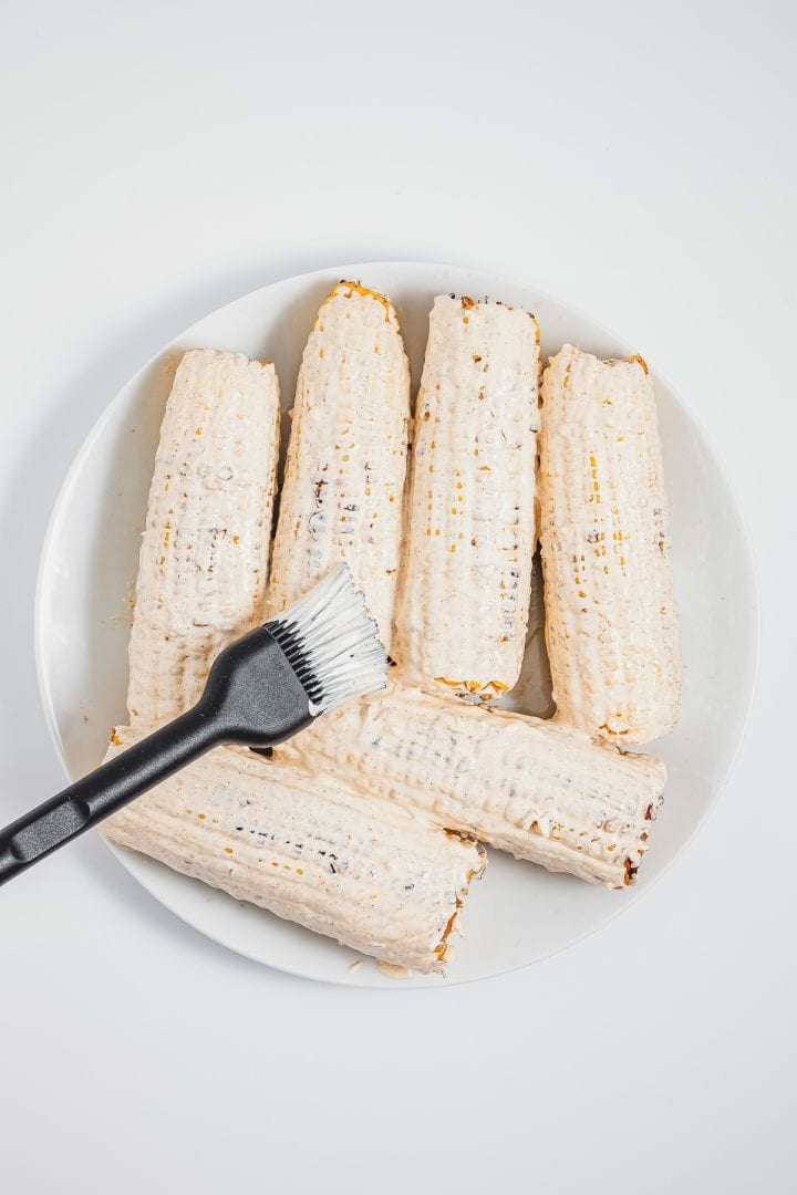 Five grilled corncobs, prepared in the style of Mexican street corn elotes, with white sauce-one being brushed with a black-handled basting brush on a white plate.