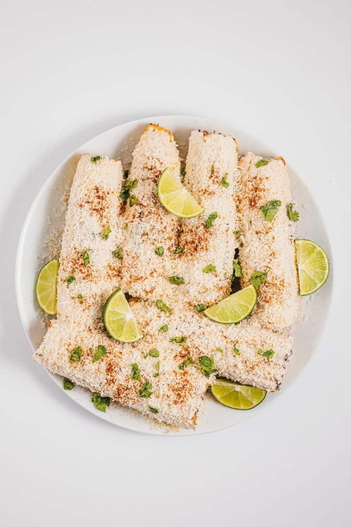 A white plate with Mexican street corn elotes, sprinkled with cheese, spices, herbs, and lime wedges on a white background.