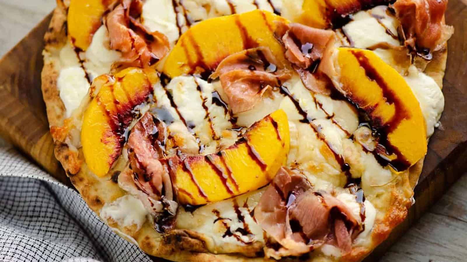 Flatbread topped with peach slices, prosciutto, ricotta, and a drizzle of balsamic glaze on a wooden board.