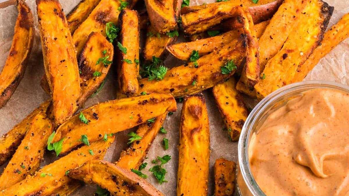 Sweet potato fries garnished with herbs, served on parchment with a bowl of creamy dipping sauce.