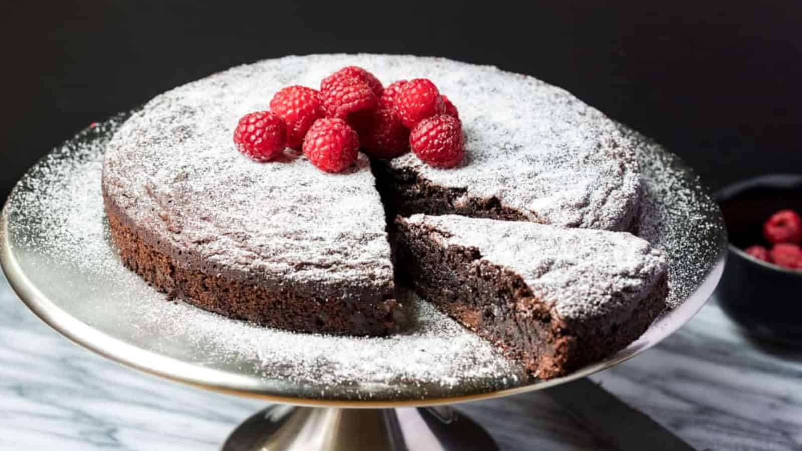 Chocolate cake topped with powdered sugar and raspberries, with a slice cut out, on a cake stand.