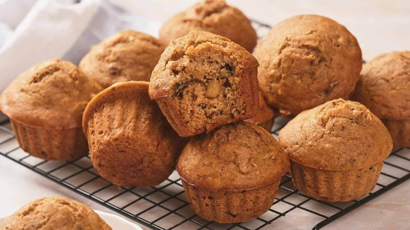 A batch of brown muffins rests on a cooling rack, with one muffin on top showing its crumb and nut pieces inside.