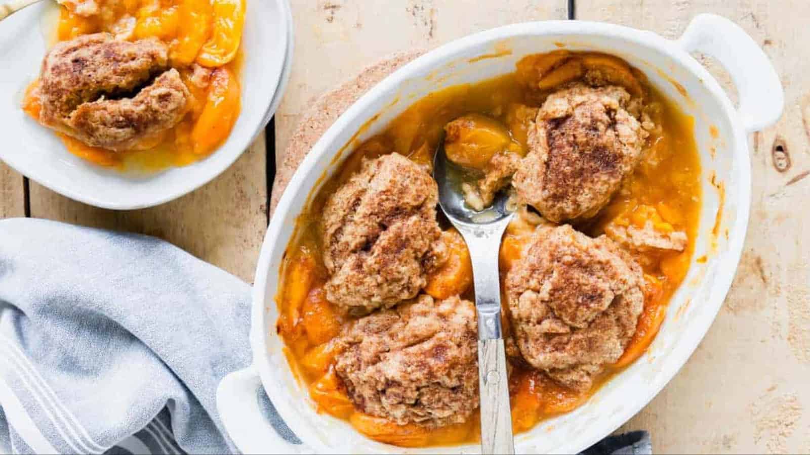Peach cobbler with cinnamon biscuits in a white baking dish, served with a portion on a white plate nearby.