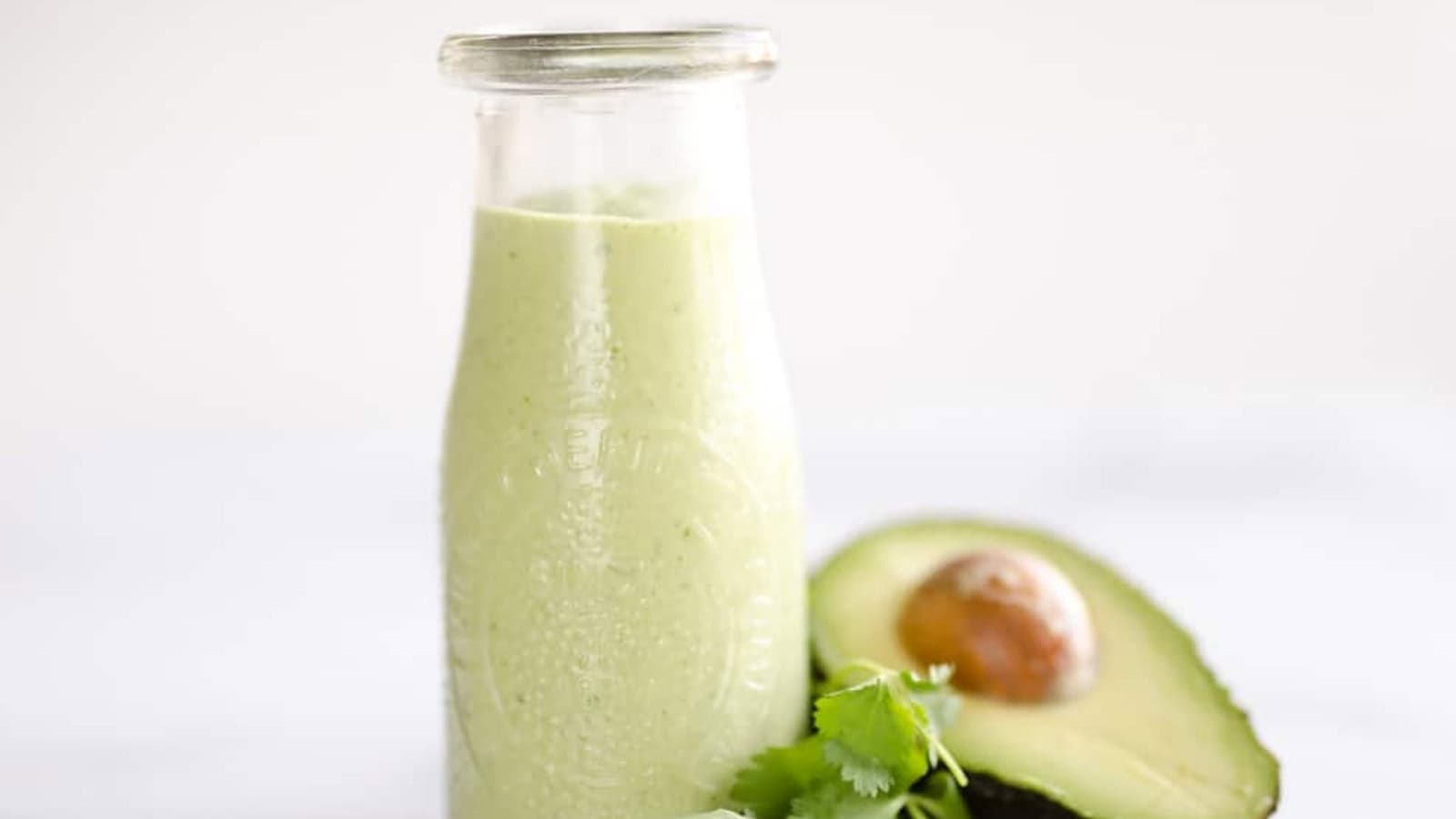 A glass bottle of green avocado dressing next to a halved avocado and a sprig of cilantro.