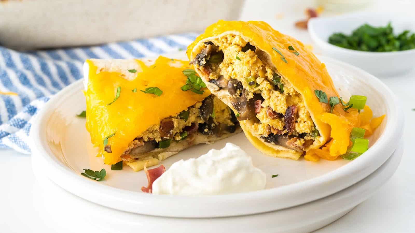 Breakfast burritos with melted cheese, eggs, mushrooms, and herbs on a plate, served with sour cream.