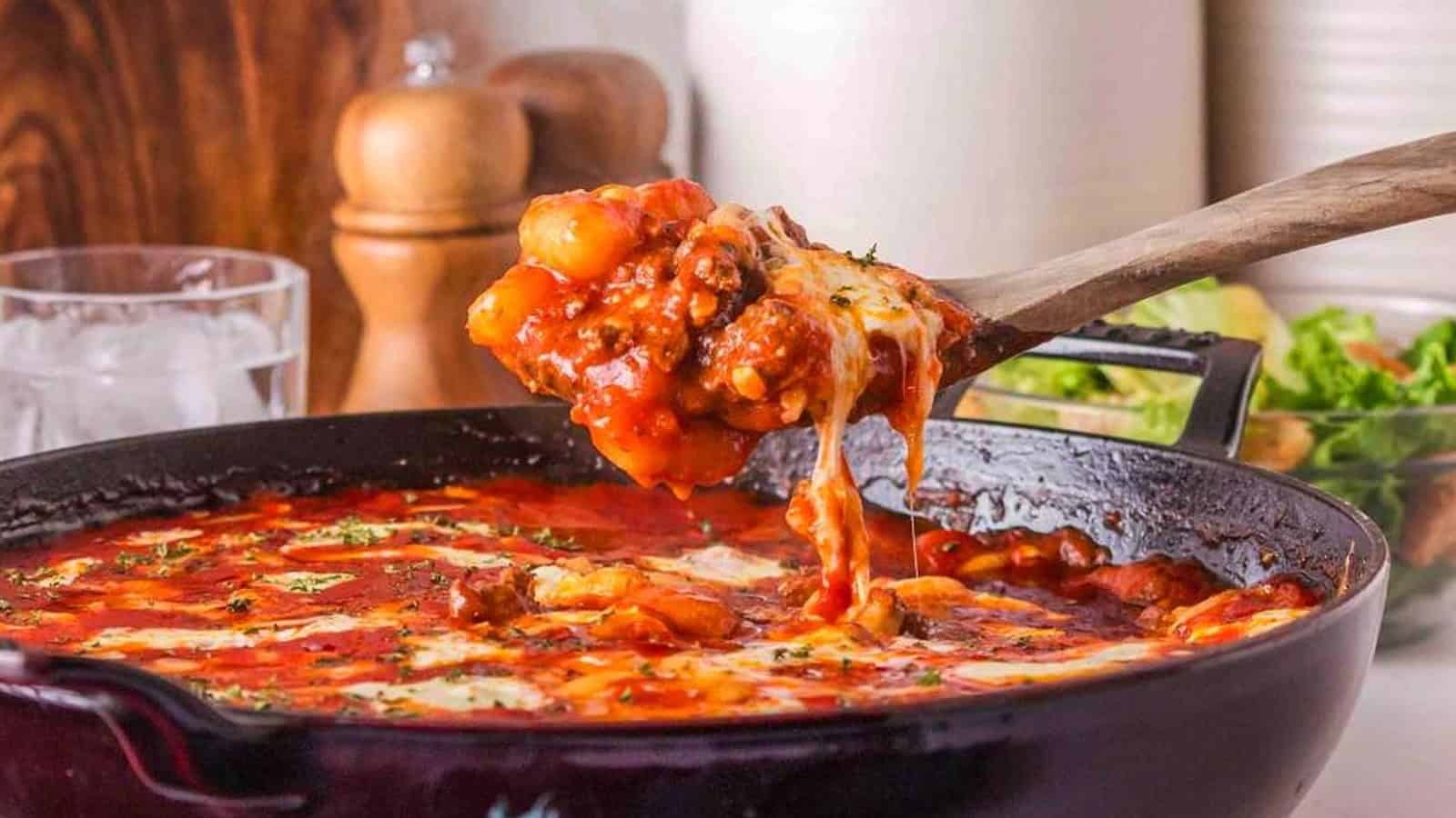 A wooden spoon lifts cheesy baked gnocchi with tomato sauce and meat from a skillet.