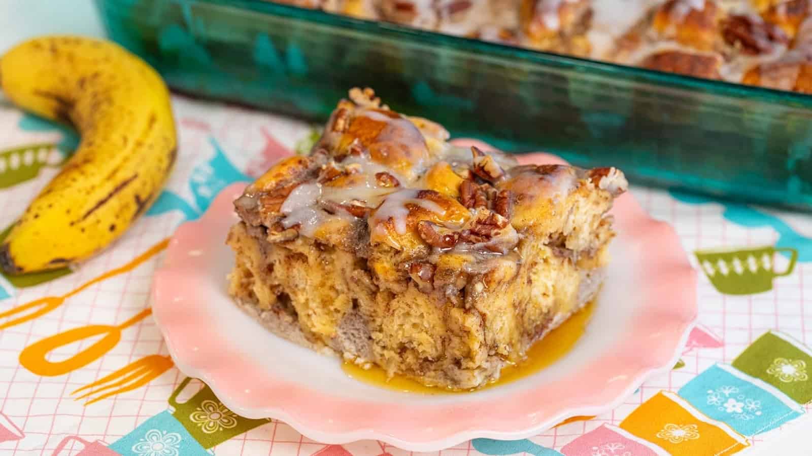 A slice of glazed bread pudding with pecans on a pink plate, next to a ripe banana.