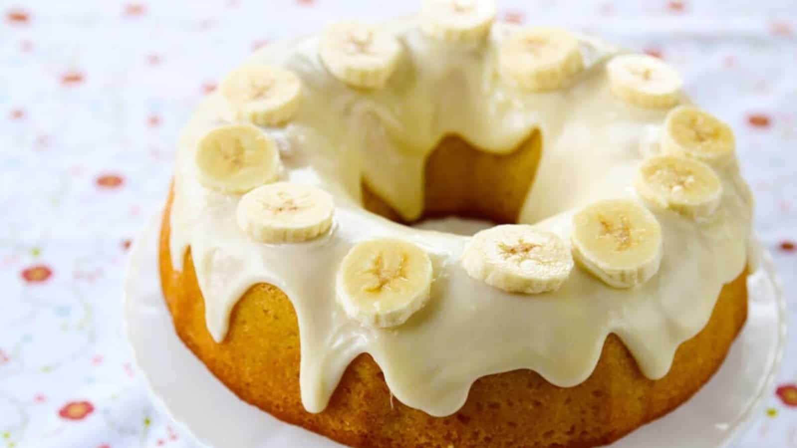 Bundt cake with white icing, topped with banana slices, on a white plate with a floral tablecloth.