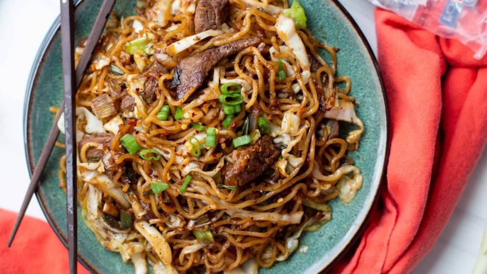 A bowl of stir-fried noodles with beef, cabbage, and green onions, served with chopsticks and a red napkin.
