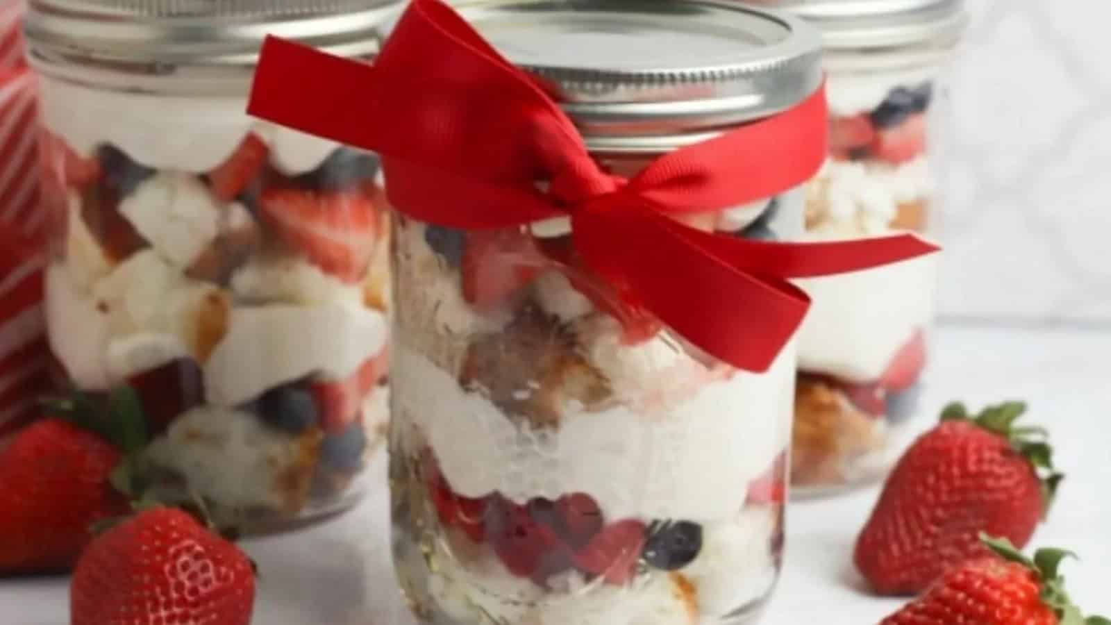 Dessert jars with whipped cream, berries, and cake, one jar tied with a red ribbon, strawberries nearby.