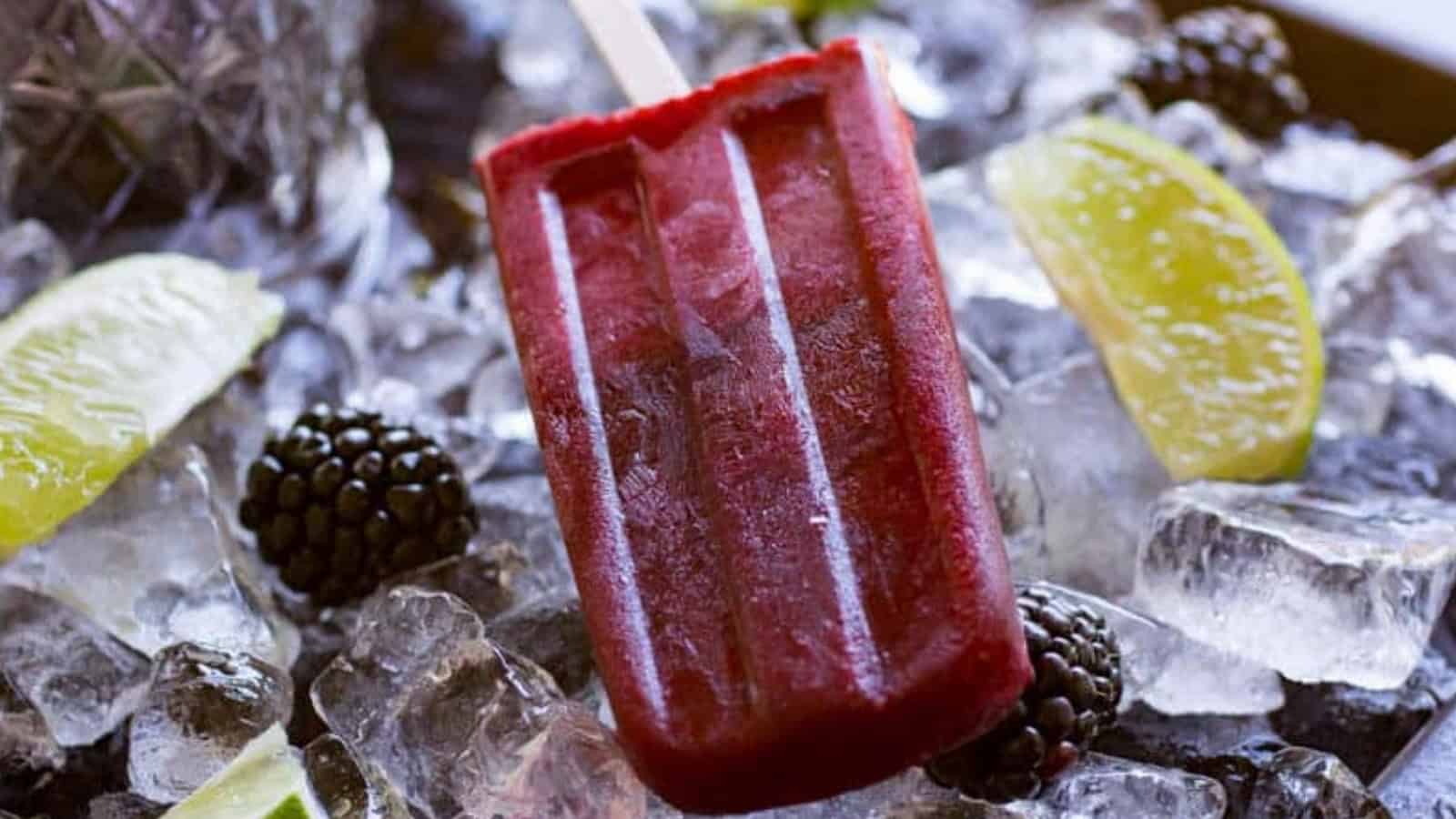 A red popsicle on ice, surrounded by blackberries and lime wedges.