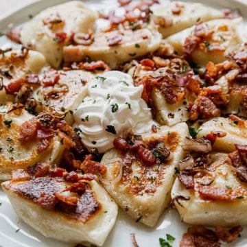 A plate of pierogies topped with crispy bacon, sautéed onions, herbs, and a dollop of sour cream.
