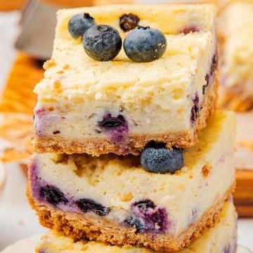 Three blueberry cheesecake bars stacked, topped with fresh blueberries, with more bars and berries in the background.
