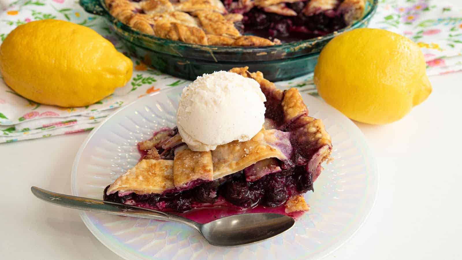 A slice of berry pie with vanilla ice cream on top, next to a spoon and two lemons.